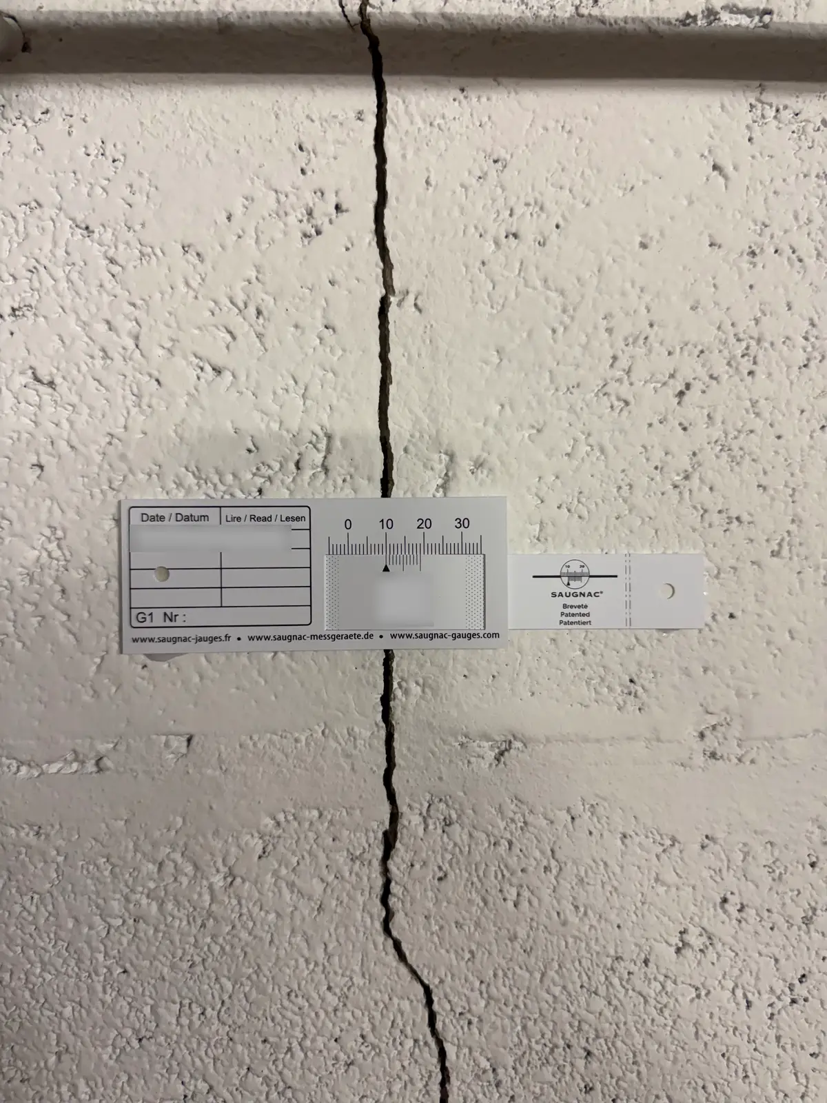 Une jauge graduée posée à cheval sur une fissure verticale dans un mur peint en blanc.