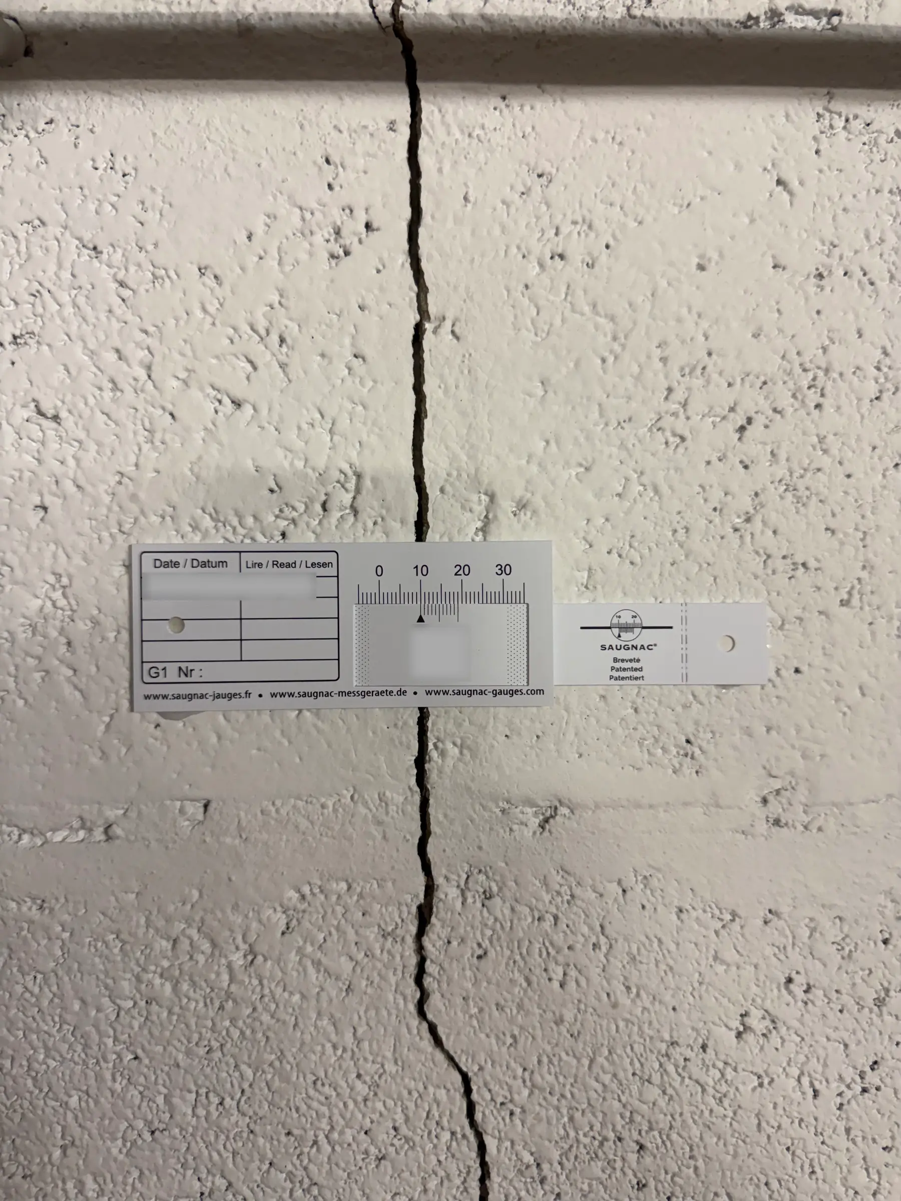 Une jauge graduée posée à cheval sur une fissure verticale dans un mur peint en blanc.